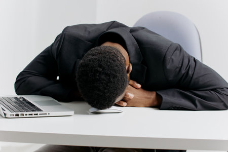 Exhausted businessman in a suit resting his head on desk amidst piles of paperwork in office settingの写真素材