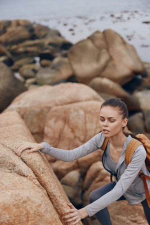 Young woman climbing rocks by the coastline, showcasing determination and outdoor adventure in a relaxed outfit with a backpackの写真素材