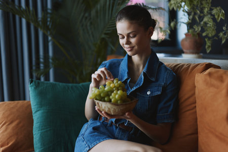Woman Enjoying a Relaxing Evening Snack on Couch with Fresh Grapes During Leisure Time at Homeの写真素材