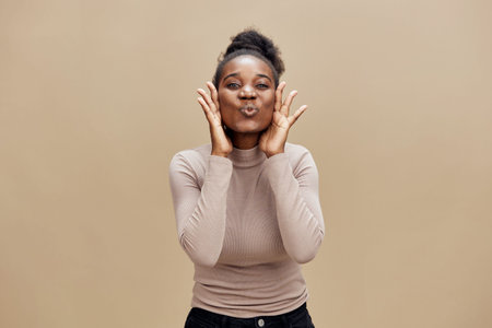 Happy African woman expressing joy with playful gestures against a warm beige background, showcasing a stylish outfit and a vibrant smileの写真素材