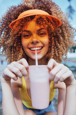Happy girl with curly hair enjoying a refreshing smoothie on a sunny day, wearing a colorful visor