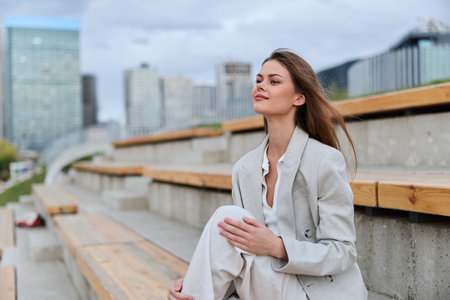 Confident woman in business attire sits on outdoor steps, reflecting on her day amidst a modern urban backdrop, exuding calm professionalism and poise.の写真素材
