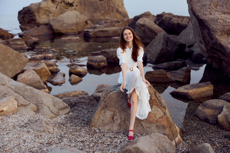 Serene woman in white dress sitting on ocean rock with rocky backdropの写真素材