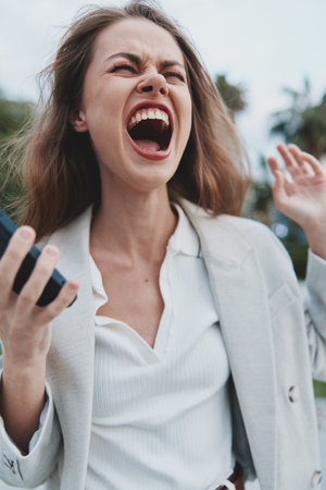 frustrated woman shouting holding smartphone outdoors expressing financial crisis emotions wearing white shirt and light blazerの写真素材