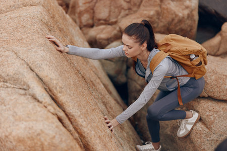 Young woman climbing rocky terrain, showcasing determination and adventure, dressed in outdoor activewear with a backpack against warm earth tonesの写真素材