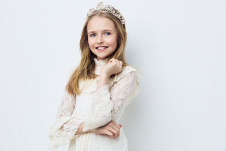 Cheerful girl wearing a crown and elegant dress, posed against a soft white background, showcasing innocence and joy, perfect for children s fashion themesの写真素材