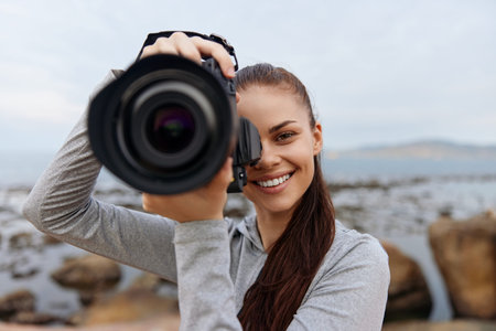 Young woman smiling and holding a camera at the beach, showcasing joy and passion for photography, with a blurred seascape backgroundの写真素材