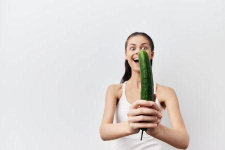 Woman holding a cucumber, smiling with excitement, casual style, light background, fresh vegetables, healthy lifestyle, fun concept, humorous pose, vibrant colorsの写真素材