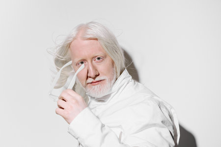 Portrait of a mature man with long white hair and a beard, wearing a white outfit, against a plain...