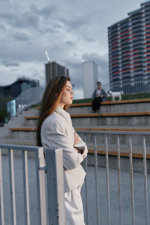 Woman in neutral coat standing thoughtfully near metal fence in urban setting with modern buildings and cloudy sky, conveying introspective mood and fashion styleの写真素材