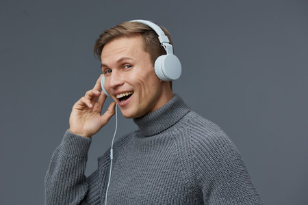 smiling cheerful young caucasian man wearing gray knitted turtleneck sweater listen music with white...