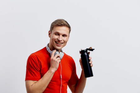 Smiling happy young caucasian man wear red t-shirt headphones hold bottle look camera isolated white background fitness healthy lifestyle conceptの写真素材