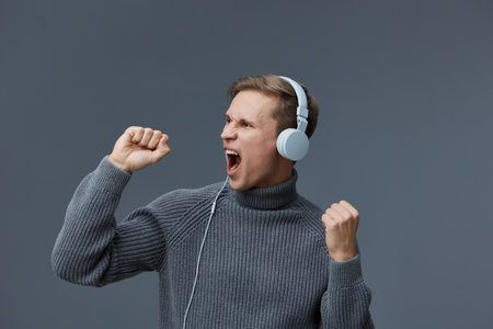 Excited young caucasian man wearing gray knitted sweater headphones celebrating success isolated on solid gray background digital content generation conceptの写真素材