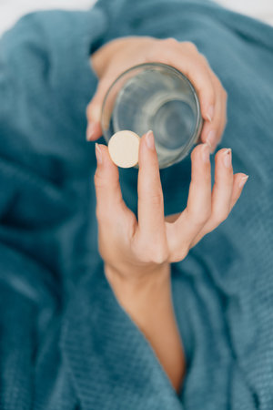 Woman in blue blanket holding glass of water with pill in hand and mouthの写真素材