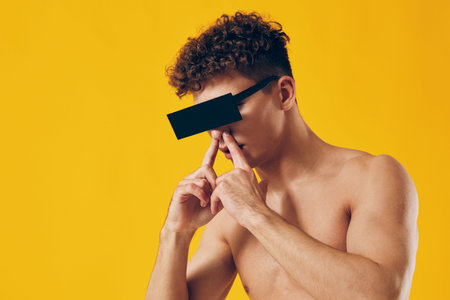 Thoughtful young man with curly hair posing on a bright yellow background, wearing sunglasses and expressing deep contemplation. Fashion and style concept.の写真素材