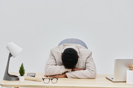 Overworked man resting his head on desk in office environment, exhausted and stressed, surrounded by workplace items like laptop, glasses, potted plant, and notepad, concept of work fatigue and burnoutの写真素材
