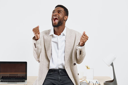 Smiling man with beard, joyful expression, wearing smart casual blazer and white shirt, standing in modern office with laptop, celebrating success, light background, positive work attitude, lifestyle concept.の写真素材