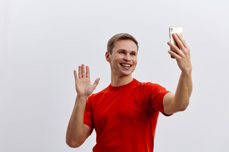 Happy man taking selfie with smartphone, smiling and waving hand, casual red t-shirt, positive emotions, indoor studio portrait with plain light background, lifestyle conceptの写真素材