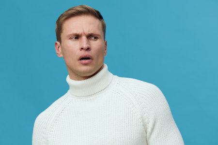 Man with confused expression wearing white turtleneck sweater isolated on blue background studio portrait. People facial expression conceptの写真素材