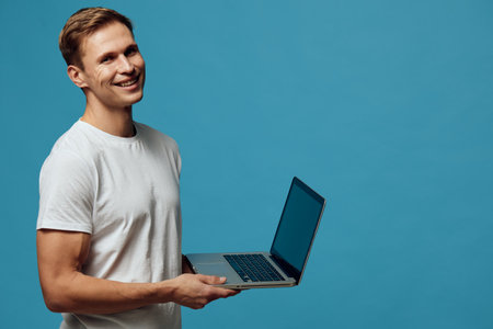 smiling young caucasian man hold laptop use remote work digital nomad concept on solid blue background casual white t-shirt technologyの写真素材