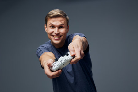 Smiling man with short brown hair wearing blue casual shirt holding game controller, looking directly at camera with cheerful expression, isolated on plain dark gray background, studio portrait, people lifestyle conceptの写真素材