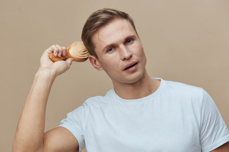 Casual young man with light brown hair holding a wooden brush near his ear, wearing a plain white t-shirt, against a neutral beige background. Studio portrait, lifestyle concept, natural expression, grooming, male hygiene.の写真素材