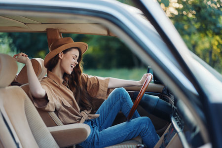 Happy young woman enjoying a road trip in a vintage car, wearing a stylish hat and relaxed outfit, with a natural green background and warm sunlight.の写真素材