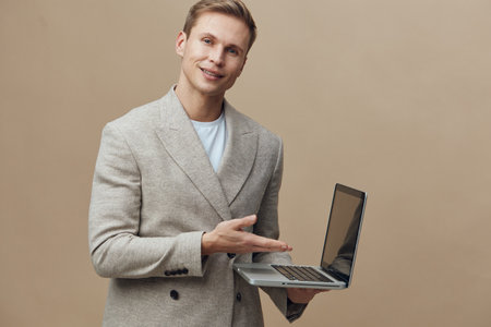 Smiling man holding laptop computer in hand looking at camera, casual business attire, indoor studio...