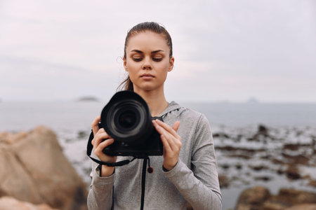 Young female photographer in a gray hoodie, focused on her camera at a serene beach with rocky shore, capturing natural beauty in soft, muted colorsの写真素材