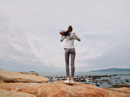 A young woman in activewear standing on rocks by the ocean, enjoying nature s tranquility and showcasing a healthy lifestyle in a serene coastal settingの写真素材
