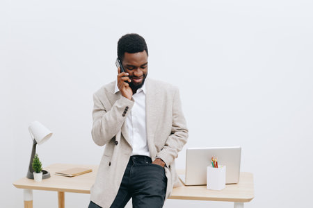 Professional young African American man in casual blazer and shirt talking on mobile phone while standing at modern workspace with laptop, desk lamp, and plants. Business communication, work, technology concept.の写真素材