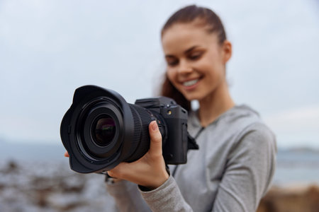 Young woman with a camera at the beach, wearing a gray hoodie, smiling joyfully towards the ocean, capturing memories in a beautiful coastal settingの写真素材