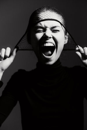 Expressive young woman in black turtleneck, dramatically pulling at fishnet headpiece while showcasing strong emotions. Bold and artistic black and white concept.の写真素材