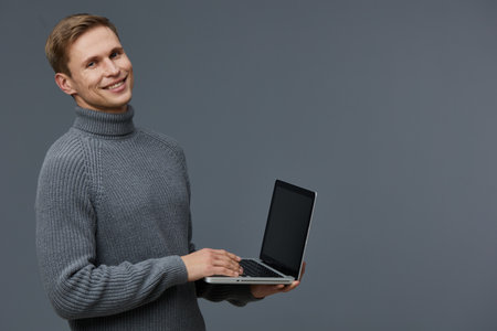 Smiling young man with light brown hair wears cozy gray turtleneck sweater and holds open black laptop, standing against plain light blue background. Casual people lifestyle concept.の写真素材