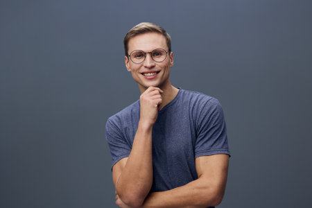 Smiling young man with glasses wearing casual blue t-shirt, posing confidently against a plain gray...