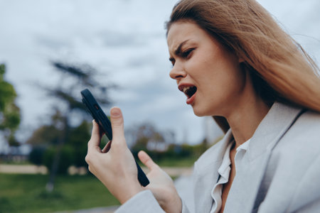 frustrated woman holding smartphone outdoors with intense emotion in casual business attire expressing financial crisis and stress in overcast urban parkの写真素材