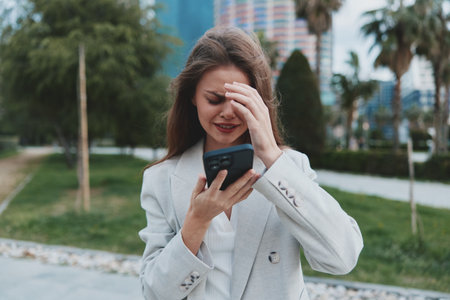 upset woman in gray blazer looking at smartphone with stressed expression outdoors in urban park, financial crisis conceptの写真素材