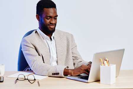 Professional smiling African American man in beige blazer working on a laptop at desk in modern...
