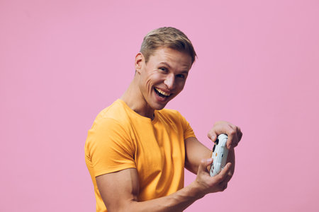 smiling young caucasian man in orange t-shirt holding gamepad enjoy digital entertainment video games isolated on pink background cheerful gamer lifestyle conceptの写真素材