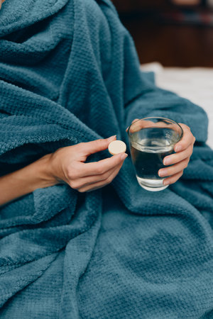 Woman wrapped in blue blanket sitting on bed holding glass of water and pill in hand