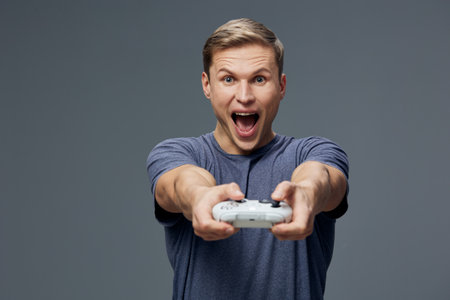 Excited young man holding game controller with open mouth, celebrating a win, wearing casual blue...