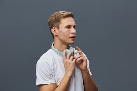 Young man with short light brown hair, wearing a white t-shirt and headphones around neck, looks...