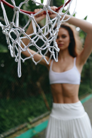 Athletic young woman in a white sports outfit with a basketball hoop, showcasing strength and confidence in a vibrant outdoor settingの写真素材