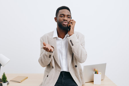 Confident young man talking on mobile phone indoors at modern workspace, wearing smart casual outfit and jacket, expression engaging, professional setting with laptop, stationery, minimal decor, bright natural lighting.の写真素材