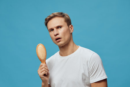Male young adult with puzzled expression holding hairbrush in hand isolated on blue background. People emotion conceptの写真素材