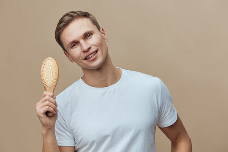 Smiling young man with short brown hair using wooden hairbrush on beige background, casual white t-shirt, positive mood, grooming, skincare, everyday routine, studio portrait, lifestyle concept, modern style, male grooming.の写真素材