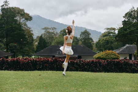 A young woman in a white sporty outfit dancing joyfully in a vibrant green park, showcasing her happiness against a scenic mountain backdropの写真素材