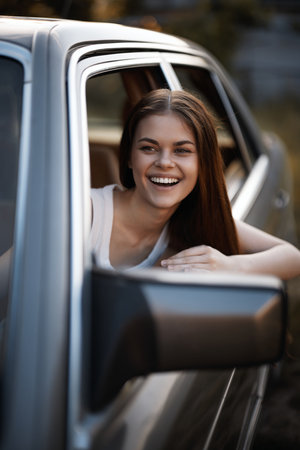 Cheerful young woman smiling from a car window, showcasing summer vibes and joy in a natural outdoor...