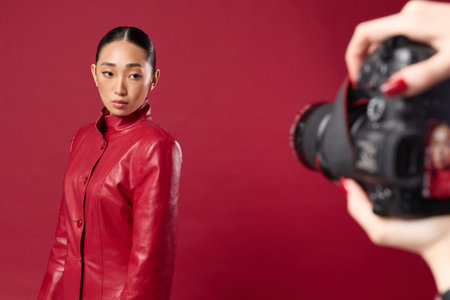 Fashion asian brunette woman in red leather jacket taking a photo of another person against red background with digital cameraの写真素材
