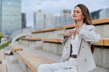 Confident woman in beige business suit sitting outdoors on urban benches with modern city background, calm expression and professional style in spring seasonの写真素材